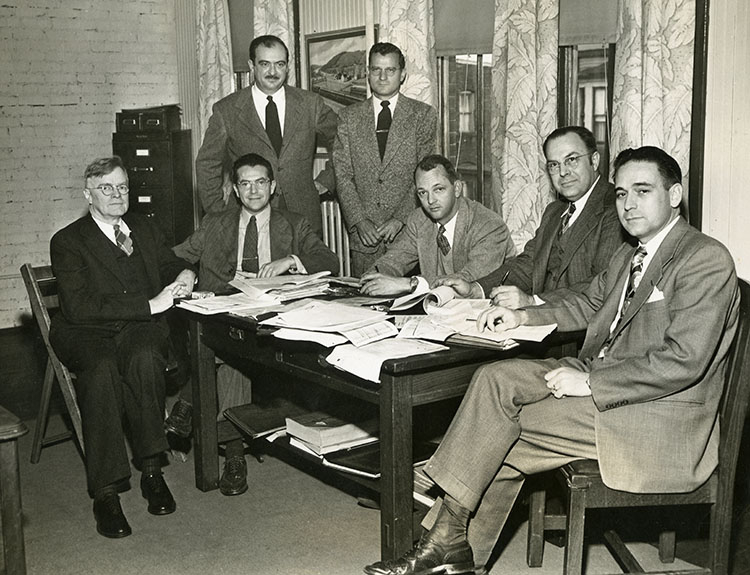 Group of 7 men in suits seated around a conference table