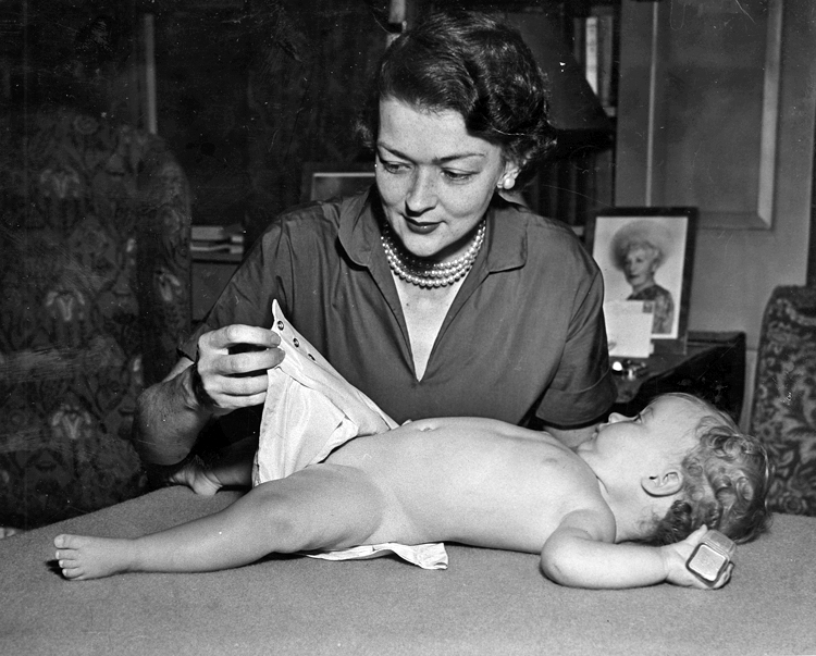 A baby holding a block in one hand lies on a table and looks up at Marion O’Brien Donovan, who is diapering the baby, using her waterproof, reusable diaper cover. She is looking down at the snaps in the cover that allow it to be adjustable to the baby’s size without the use of safety pins. She is smiling slightly and is wearing a V-neck dress and a triple strand of pearls. A framed photo of an older woman wearing a large hat is visible on a table over Donovan’s left shoulder.