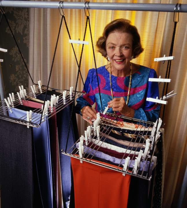 Marion O’Brien Donovan smiles at the camera. She is standing behind 2 Big Hang-Up organizers and is placing a clothespin on one garment to attach it to the rack. A backlit beige curtain is behind her and the organizers hang from a horizontal metal pole. A few clothespins are attached to the cords that suspend the organizer from the pole.