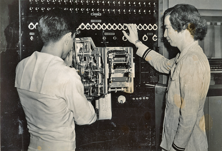 A young navy ensign stands with his back mostly to the camera and next to Grace Hopper, wearing her navy uniform and standing in profile to the camera. She has her right hand on one of the many dials that run across the top of the Mark I computer. She and the ensign are looking at a section of the computer with many gears to move the punched tape code through the machine.