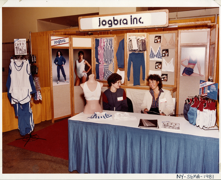 Hinda Miller (left) and Lisa Lindahl sit at a skirted trade show table in 1981. The 2 women are looking at each other and smiling. A half-torso mannequin is on the table, modeling a Jogbra. Other Jogbra Inc. merchandise, including gloves, track suits, bras, shorts, and one-piece running outfits, is displayed on the table and behind them on the booth walls, along with large photographs of women wearing some of the gear.