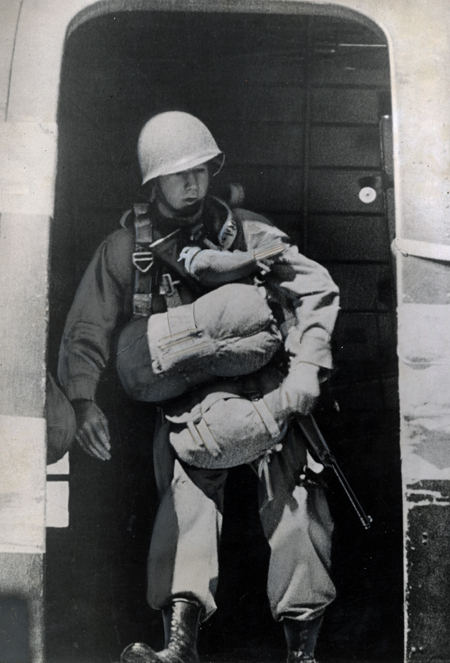 A paratrooper, carrying full gear and a gun and wearing a helmet, stands at the open hatch with a pigeon attached to his chest. The pigeon wears a specially-designed vest to protect it when the paratrooper jumps from the plane.