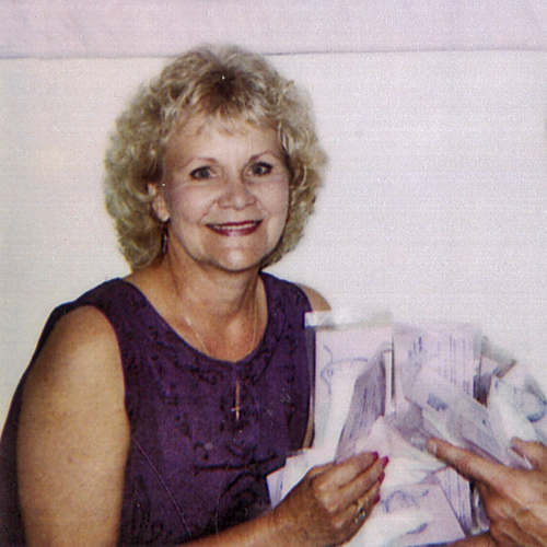 Sharon Rogone, smiling at the camera. She is holding an assortment of unidentified papers. She has curly blonde hair and is wearing a sleeveless navt blue dress. This image has been cropped from a larger photo depicting her and her business partner Kenneth Croteau.