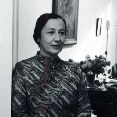 Charlotte Cramer Sachs stands in an interior doorway. A vase of flowers and paintings on the wall are visible over her left shoulder. Her hair is pulled back tightly into a bun and she wears an abstract print dress comprised of wavy diagonal lines. The photo is black-and-white.