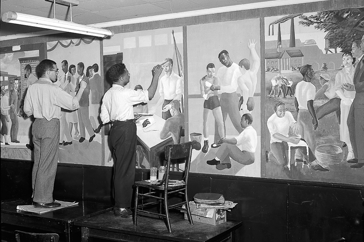 Two Black men standing on tables painting murals of Black men and boys at various activities.