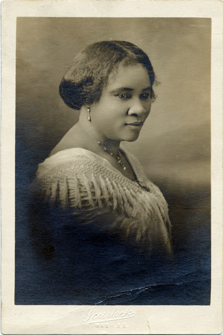 A sepia-toned 3/4 profile studio portrait photograph of Sarah Breedlove Walker, known as Madam C. J. Walker. She is wearing a shawl with tassels adorning the neckline, fastened with a brooch, and she wears drop earrings with matching necklace and a brooch.