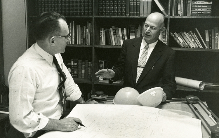 A man wearing glasses holds a pen over a blueprint drawing while Earl Tupper gestures with 2 Tupperware bowls