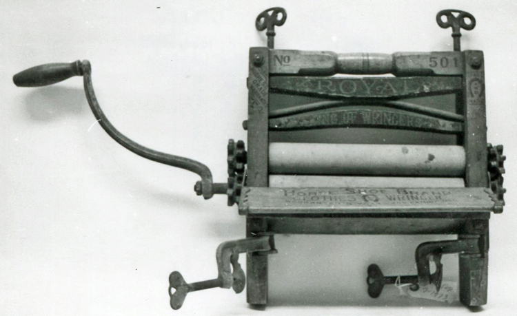 Black-and-white photo of a clothes wringer, comprised of two rubber rollers in a wood frame with metal gears, clamps, etc.