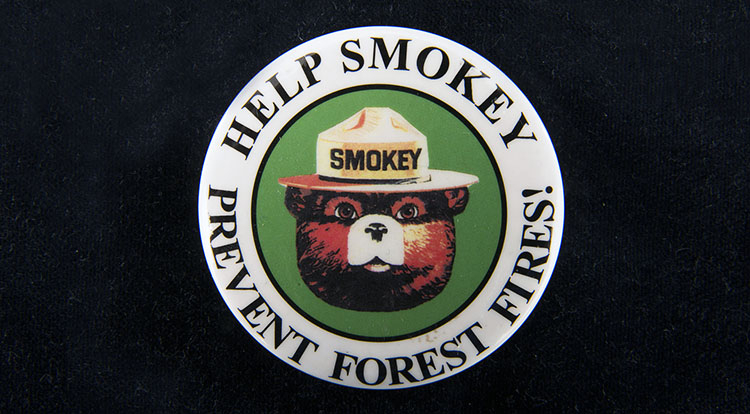 Metal button with image of Smokey the Bear’s head and the words “Help Smokey Prevent Forest Fires” around the outer edge