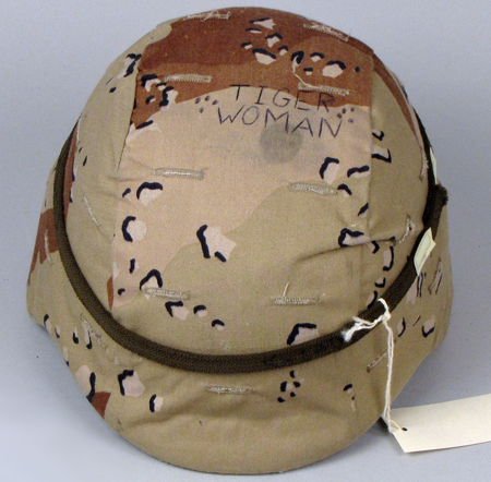 A 3/4 view from overhead of a Kevlar army helmet. The helmet is covered in tan, green, and brown camouflage-patterned cloth and the words “Tiger Woman” have been written in all capital letters on the top of the helment.