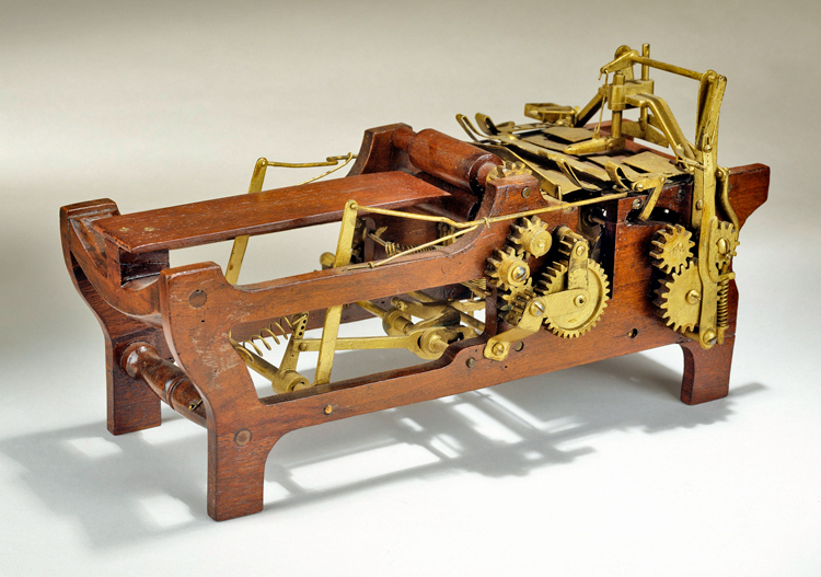 Margaret Knight’s patent model for an improved paper bag making machine shows 2 sets of 3 gears attached by articulated arms and springs to a wooden frame and horizontal bed where the paper is moved through the bag making process.