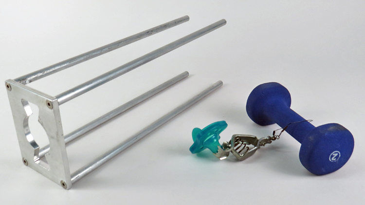 A metal platform attached to 4 spindly legs lays on its side on a table. To the right is a green pacifier attached by wire to a 5-pound blue barbell. In use, the pacifier would be placed above the opening in the stand’s platform, with the barbell suspended below. If the weight of the barbell caused the pacifier to fall through the opening, it was deemed a choking hazard.