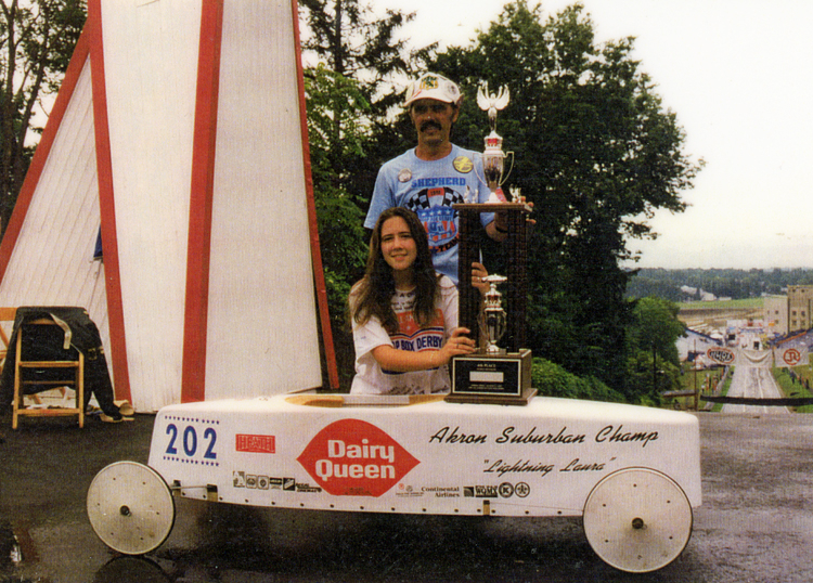 14-year-old Laura Shepherd kneels behind her soapbox derby car, holding her trophy, which is resting on the car. Her father stands behind her, with one hand on the trophy. They are at the top of a hill and the soapbox derby downhill course is visible at the right.