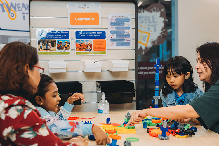 Two young inventors and two grown-ups work together at a table with an assortment of brightly colored plastic gears.