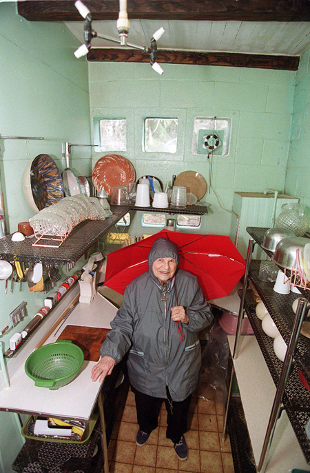 View looking down on Frances Gabe, wearing rain gear and holding an open umbrella, in her self-cleaning house kitchen in 2002