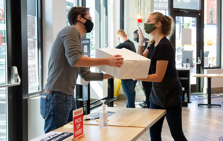 A woman handing a package of pizza to a man; both are wearing masks.