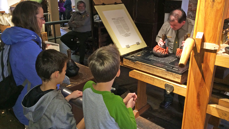 Emma, Gavin, and Patrick Hintz observe National Park Service printing demonstration on a replica of the Franklin printing press