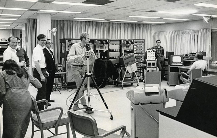 A candid photo of a group of 7 men and 1 woman in a large room filled with computer equipment. A space has been cleared in the center of the room and one of the men, Stewart Brand, is handling a camera on a tripod. 2 studio lights on stands are positioned at opposite sides of the room.