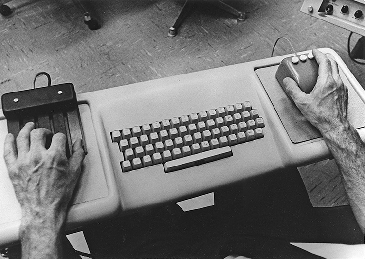 A close-up of Douglas Engelbart’s hands. He has his left hand on the NLS 5-button chord keyset, and his right hand holds a 3-button mouse; between the keyset and the mouse is a standard QWERTY keyboard. Platforms for the mouse and keyset are integrated into the keyboard base.