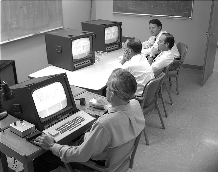 4 men sit at 3 computer terminals. Douglas Engelbart, wearing a wired earpiece, is closest to the camera and is seen in profile. His terminal has a keyboard and he has his hand on a prototype mouse. His computer is linked to the 2 terminals that the other 3 men are watching. Bill English is farthest from the camera.