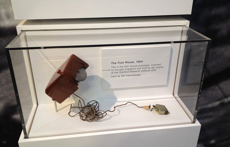 A prototype of the first computer mouse sits on an angled bracket inside a glass display case. The mouse is a dark wooden box with a red button on top. On the underside is a vertical metal wheel. A thin computer cable extends from the back of the mouse.