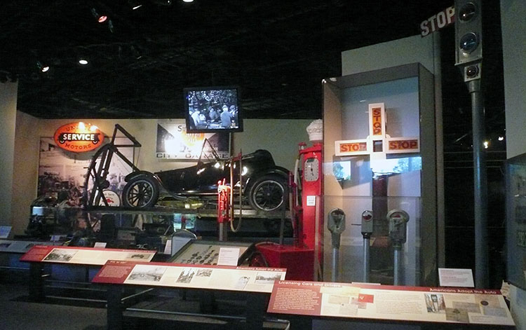 View into part of the exhibition. A car, service station sign, gas pump, parking meters, and Morgan’s traffic signal are visible.