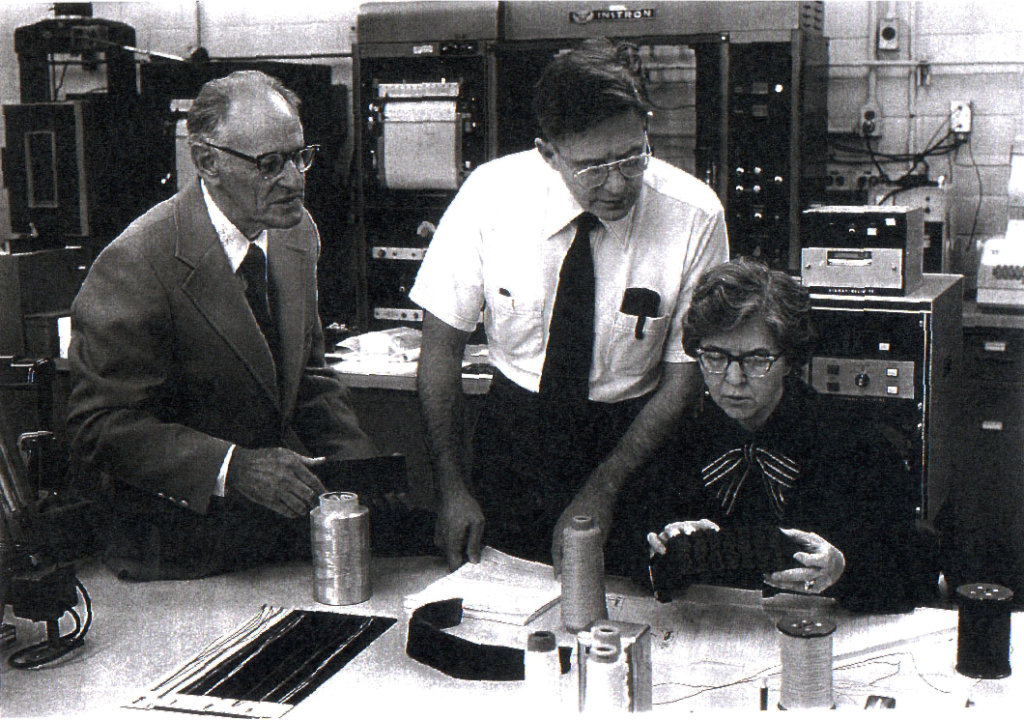 Dr. Paul Morgan, Dr. Herbert Blades, and Stephanie Kwolek are pictured in the DuPont Textile Fibers Pioneering Research Laboratory. Kwolek sits at a table holding a Kevlar sample while the men look on. 1960s-era computer equipment is in the background.