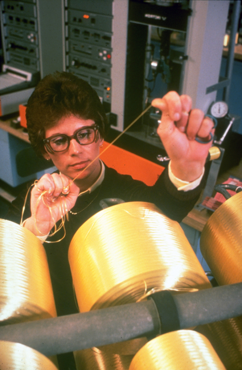 A woman draws a strand of Kevlar from one of several spools in front of her at a manufacturing site in Richmond, Virginia. 1970s-era computer equipment is in the background.