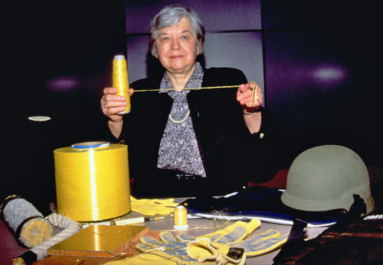 Stephanie Kwolek holds a spool of yellow Kevlar thread in her right hand and has pulled out a thread and stretched it about 16 inches with her left hand. She has short gray hair and is wearing a gray patterned shirt and pearl necklace under a black jacket. A larger spool of Kevlar fiber, a rope, gloves, and a helmet are displayed on a table in front of her.