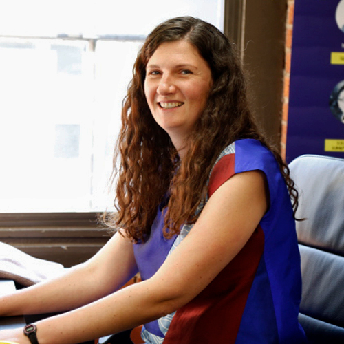 Theresa Dankovich sits in profile at her desk. She is smiling at the camera and wearing a sleeveless, blut-red-and gray top. She has long, dark, curly hair.