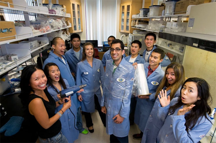 Khine with a group of students in blue lab coats pretending to shrink wrap one student