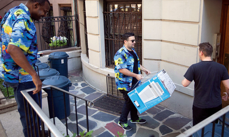 Inventor Garthen Leslie at top of stairs to a basement apartment, with Quirky founder Ben Kaufman at bottom, carrying an Aros air conditioner to be delivered. Garthen and Kaufman wear Hawaiian shirts. A third man is at the apartment building door.