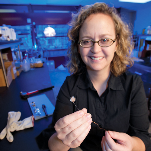 Amy Prieto in her lab at Colorado State University. She is smiling at the camera and holding a tiny prototype battery with long wire leads. Assorted chemical experimentation equipment is visible in the background. She has shoulder length, curly, sandy blonde hair and wears glasses.