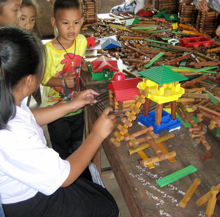 4 young children at a table covered with modern day Lincoln Logs. One girl is building a house on stilts with a ladder entrance and another traditional building surrounded by a fence, while the other children look on.