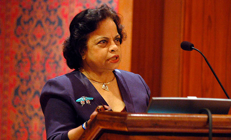 A woman speaking at a podium. She is wearing a purple suit, a silver necklace, and a large brooch of a butterfly or moth.