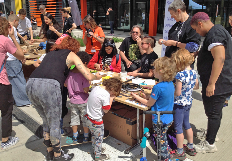 Groups of kids and their adults work at hands on projects at an outdoor table.