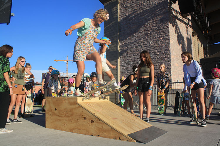 Woman on a skateboard, holding the hand of a child on a skateboard, both atop a short wooden skate ramp, about to do a trick