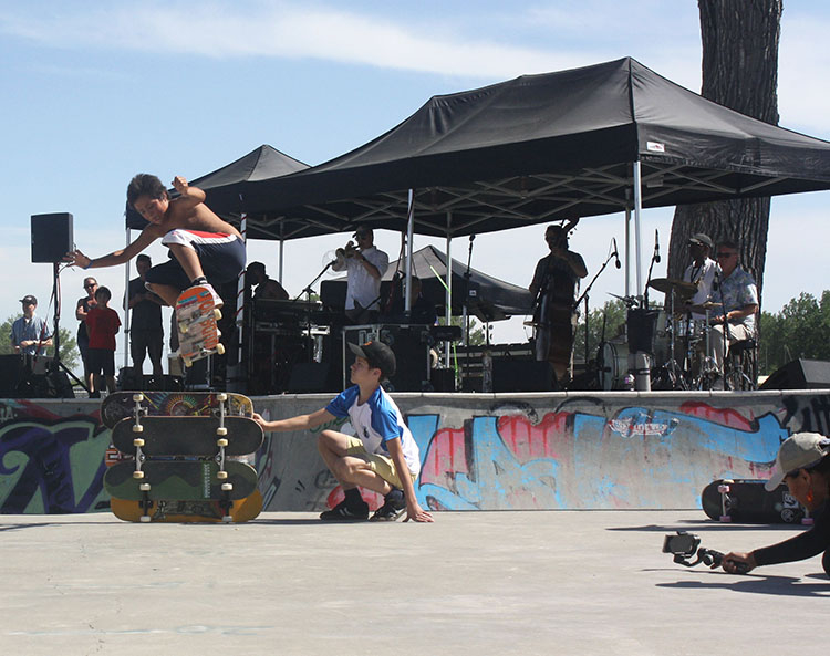 Young Lakota boy in mid-air clearing 5 skateboards stacked on horizontal edge while jazz ensemble plays behind him