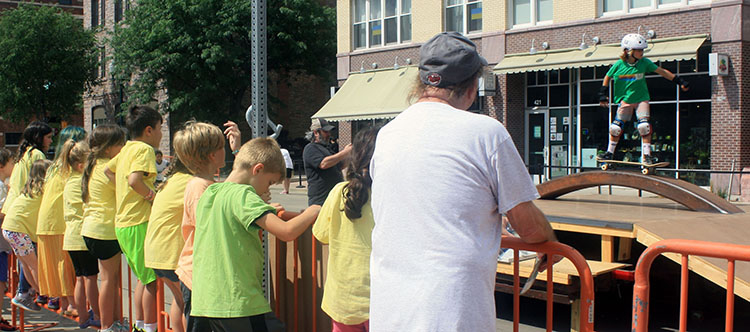 Group of people, mostly children, watch a young skater perform on street-turned-skatepark