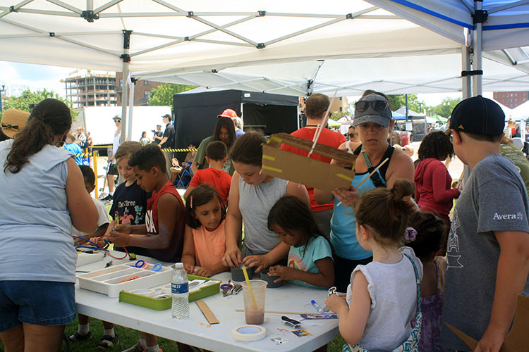 A group of children and adults work at hands-on invention activity tables under a tent canopy