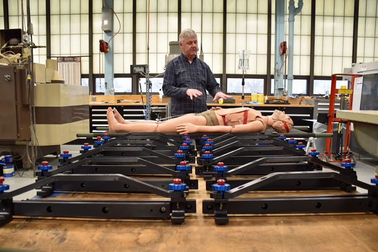 A man points to a mannequin simulating a medical patient lying on a stretcher. A rail, called the SHRAIL, is attached to the side of the stretcher and holds an IV bag. Several more unattached SHRAILs fill the foreground.