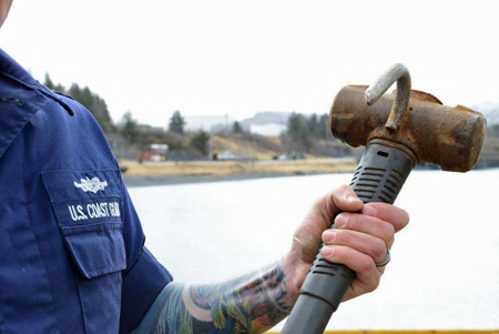 The left shoulder and tattooed arm of a man wearing a Coast Guard work shirt are visible. He holds a maul with a large hook attached to the head.