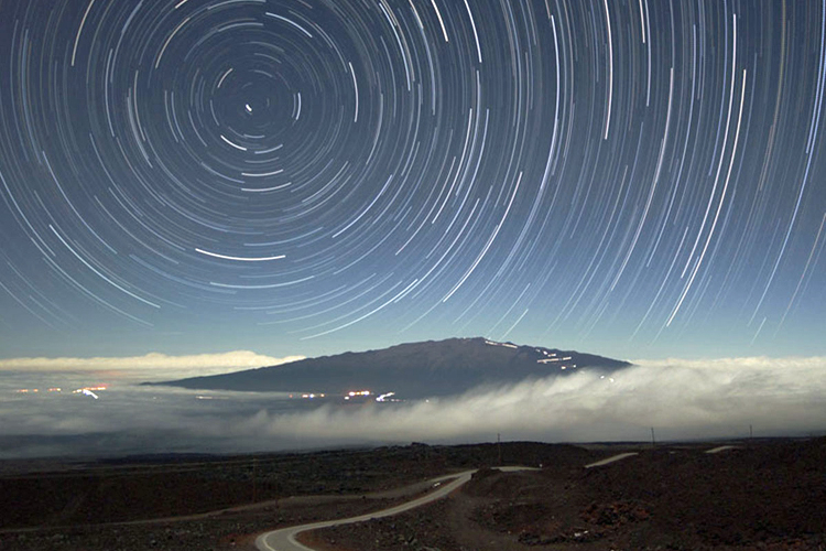 A landscape scene, showing a curving road that winds from the foreground to the background, passing under low-lying clouds, and heading toward a mountain. City lights are at the right of the image and the sky is filled with circular star trails.