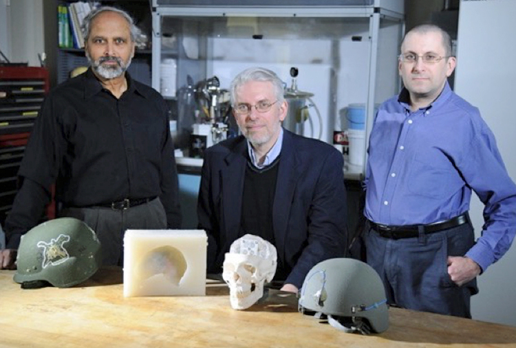 Three scientists posing with GelMan synthetic substitutes for live animals or clay materials used in testing new body armor, helmets, and flak jackets. A mold, partial skull, and 2 helmets are on the table in front of the scientists.