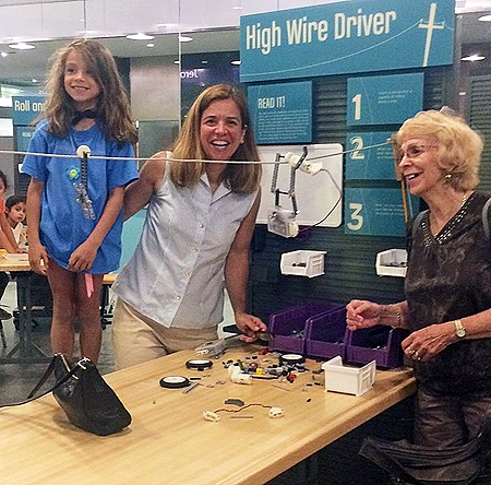 One older adult woman, one adult woman and one female child are gathered around an activity table and smiling. The picture is taken from the side view. In the middle of the picture is a light wooden table. On the table are lego pieces and wire is strung a