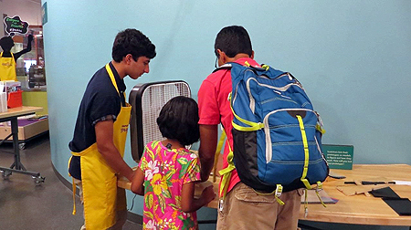 A teenage male wearing a bright yellow apron, a grown-up male, and small child are looking at a table with wooden and plastic materials on it. Their backs are toward the viewer and what they are looking at is out of sight.