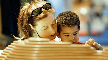 A female grown-up is sitting close to a small child. They both appear to be looking at the same thing. The grown-up is helping the child touch or explore an object that is out of sight.