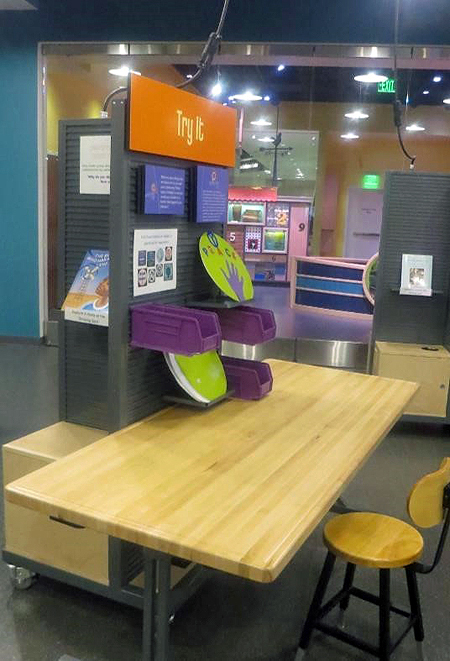 Side view of an activity station comprising a stool and a light wooden table with an attached gray panel holding 3 purple plastic buckets, 2 green circles on gray shelves, 2 purple text panels, and a large orange text panel that reads Try It.