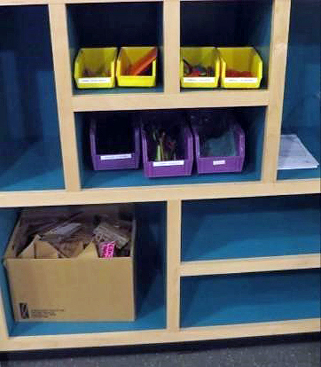 Close-up of shelves with blue interiors. On the shelves are yellow and purple plastic bins with craft supplies in them. On the bottom shelf is a cardboard box with pieces of cardboard in the box.