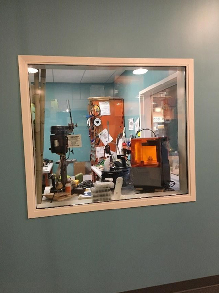 A blue wall with a clear window. Looking through the window, you can see a workshop. There are tools, 3D printers on a workbench, an orange cabinet on the right side, and pieces of paper hanging on the walls.  